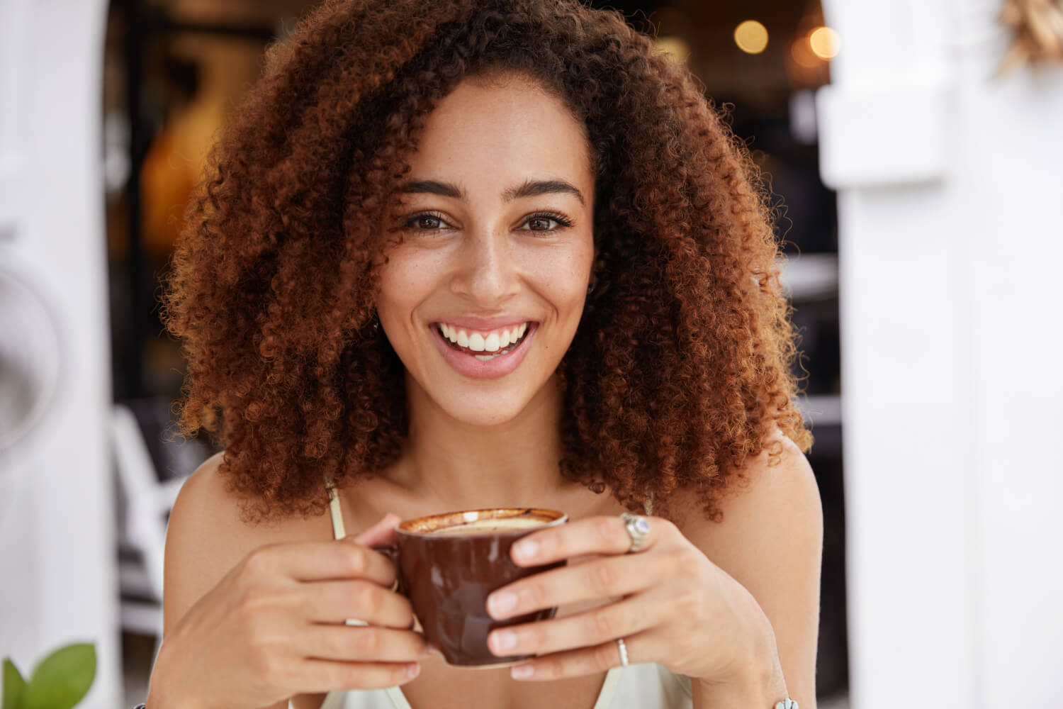 Mulher sorridente de cabelos cacheados segurando uma xícara de café. Prótese protocolo resistente a manchas na Myai Aricanduva - SP.