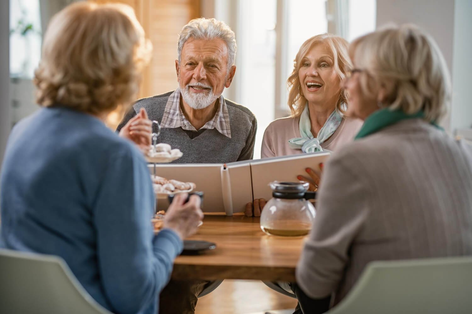 Homem idoso sorridente de barba branca conversa com três amigas em mesa, destacando reabilitação oral na Myai Odontologia no Aricanduva.